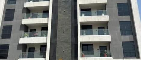 A multi-story building with balconies, a central vertical feature, and a parking area with a truck.