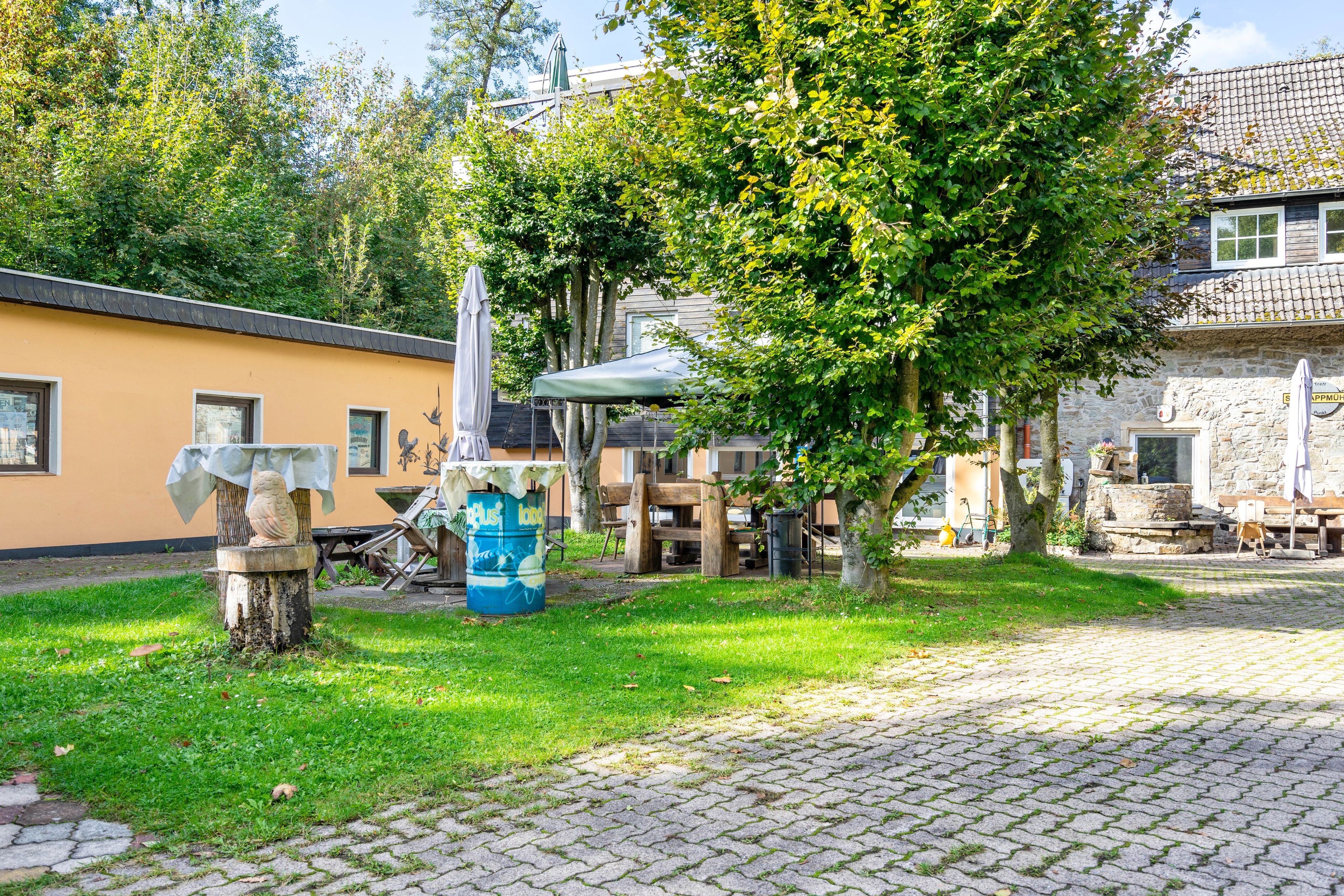 Ein Innenhof mit gepflastertem Weg, einer Holzbank, einem Tisch mit Stühlen und einem Baum, der Schatten spendet.