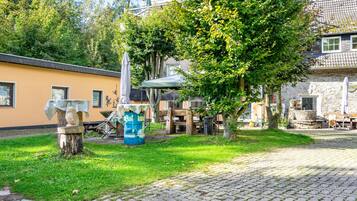 Ein Innenhof mit gepflastertem Weg, einer Holzbank, einem Tisch mit Stühlen und einem Baum, der Schatten spendet.