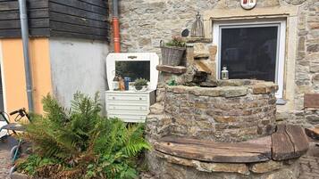 Een stenen gebouw met een houten bankje, een kleine tuin met varens en een witte dressoir met een plantenmand.
