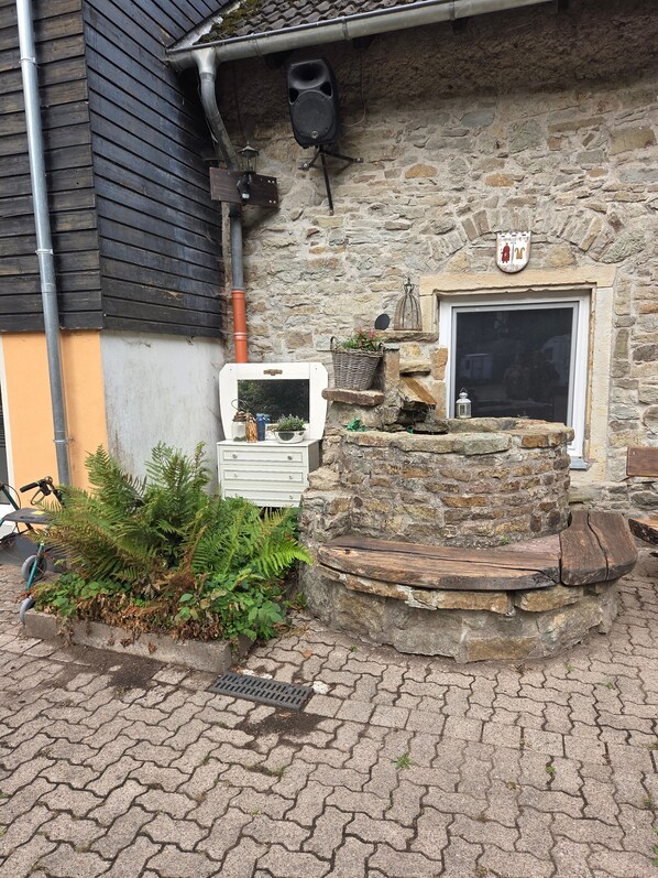 A stone building with a wooden bench, a small garden with ferns, and a white dresser with a basket of plants.