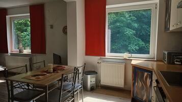 Een keuken met een eethoek, een radiator, een raam met uitzicht op bomen en een afvalbak.