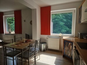 A kitchen with a dining area, a radiator, a window with a view of trees, and a trash bin.