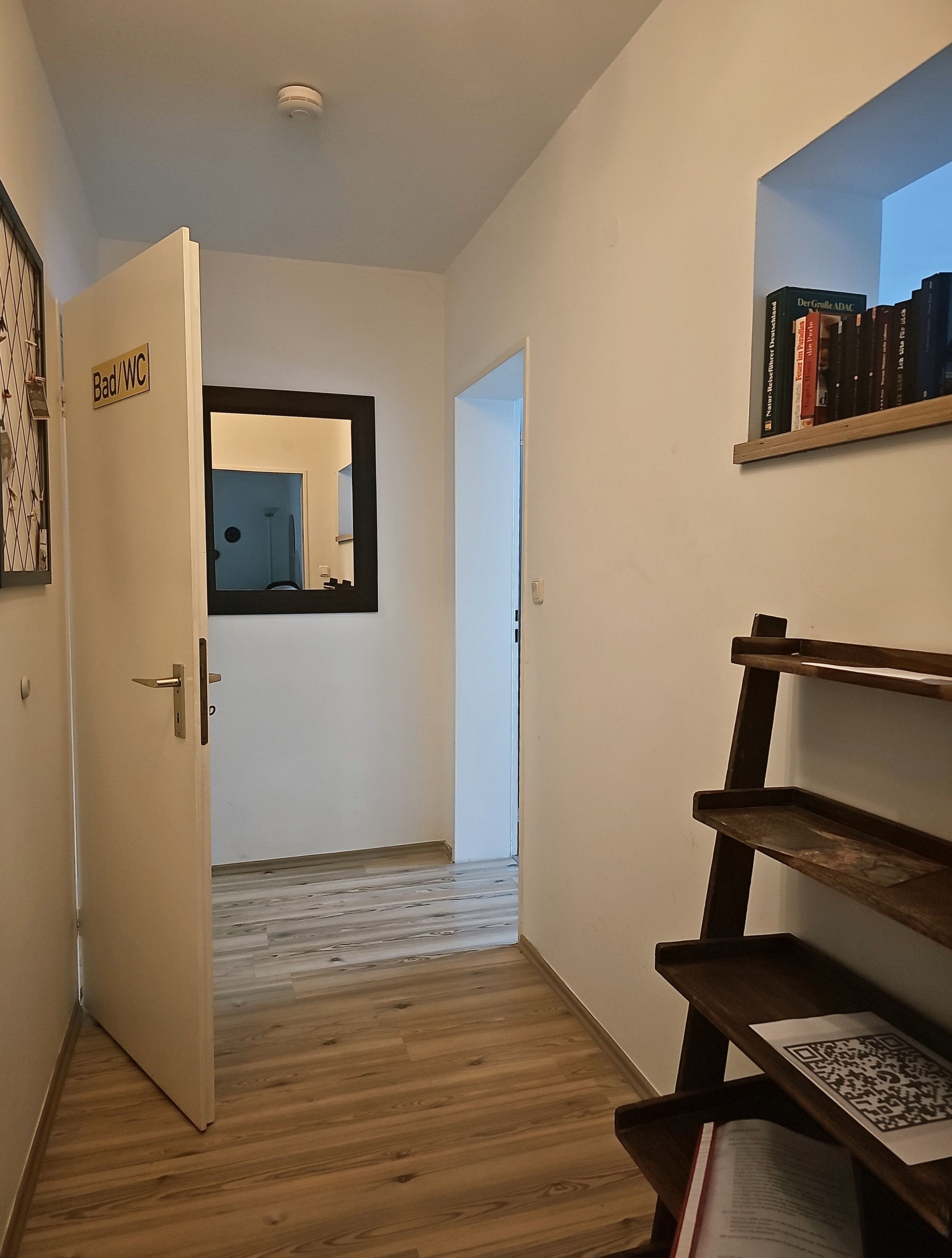 Een gang met een deur waarop 'Bad/WC' staat, die leidt naar een badkamer, een houten vloer en een boekenkast met boeken en een QR-code.