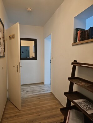 A hallway with a door labeled 'Bad/WC' leading to a bathroom, a wooden floor, and a bookshelf with books and a QR code.