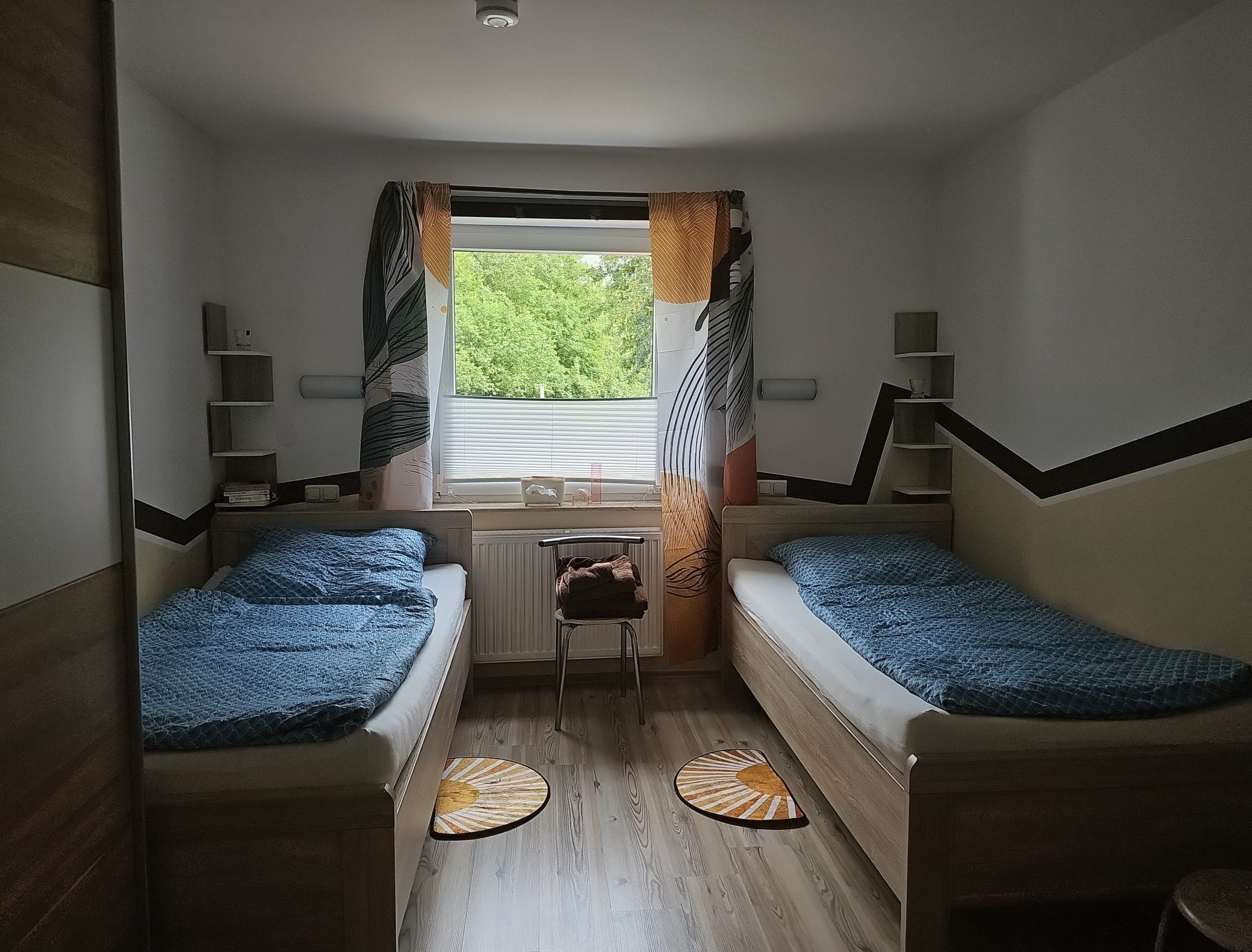 Een kleine kamer met twee éénpersoonsbedden, een bureau, een radiator en een raam met gordijnen.