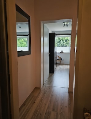 A hallway with a wooden floor leading to a room with a window, a door, and a small table with a chair.