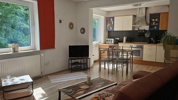 En moderne stue med sofa, sofabord og fjernsyn. Ved siden af er et køkken med spiseplads, komfur og skabe.