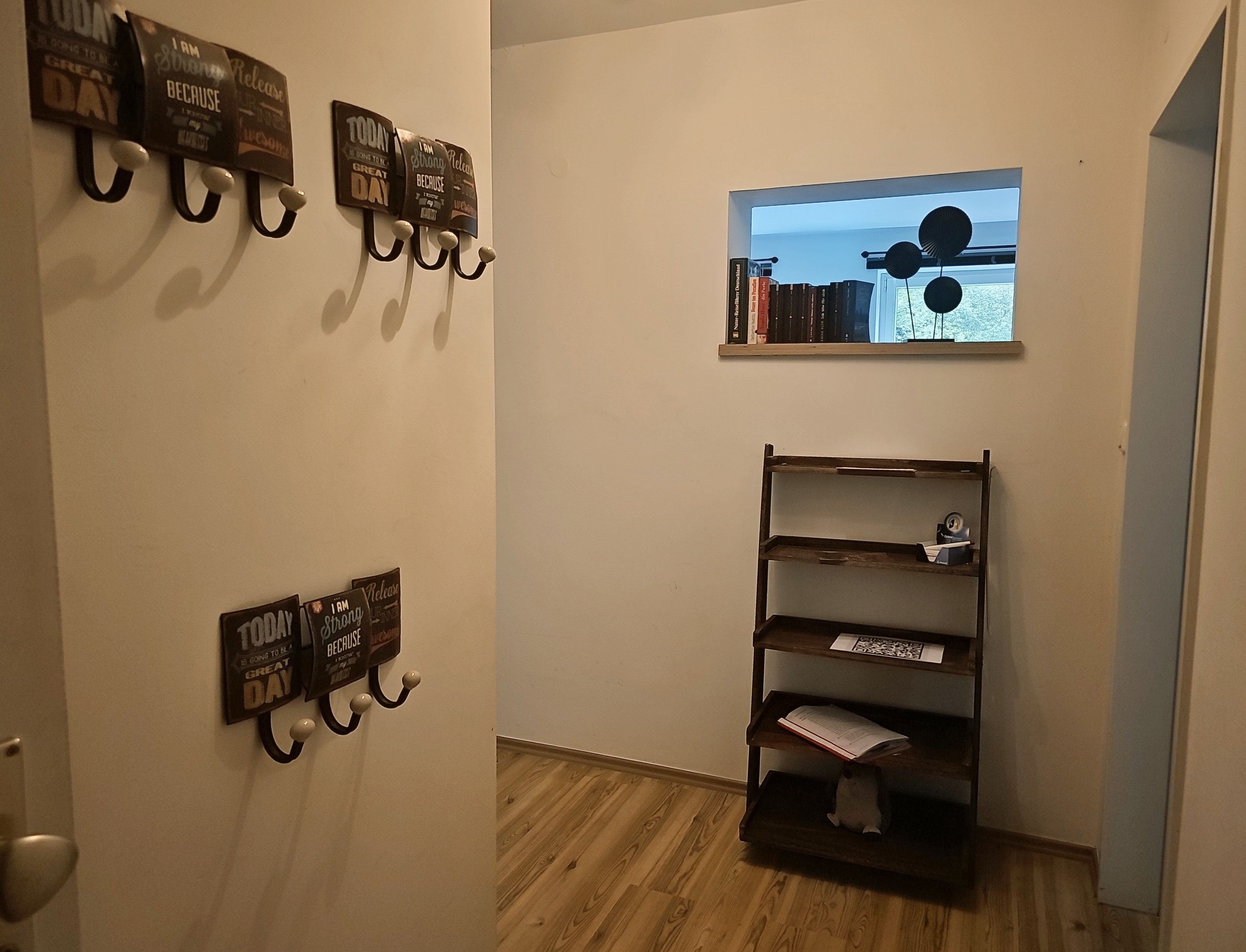 Een gang met houten vloer, een boekenplank vol boeken en wandhaakjes met inspirerende citaten.