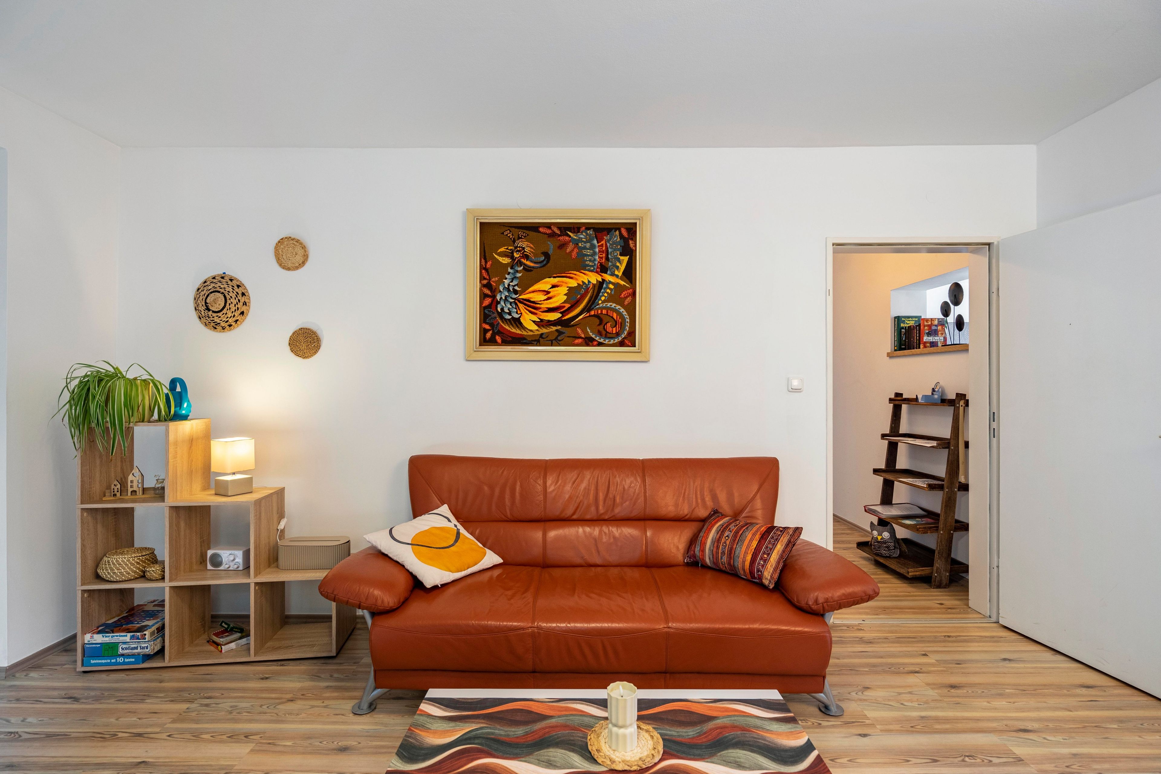 A modern living room with a brown leather sofa, a wooden side table with a lamp, a framed artwork above the sofa, and a bookshelf in the background.