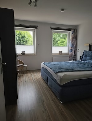 A bedroom with a bed, two windows with curtains, a chair, and a small table.