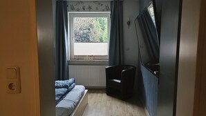 Una habitación pequeña con cama, una silla, un radiador y una ventana con cortinas.