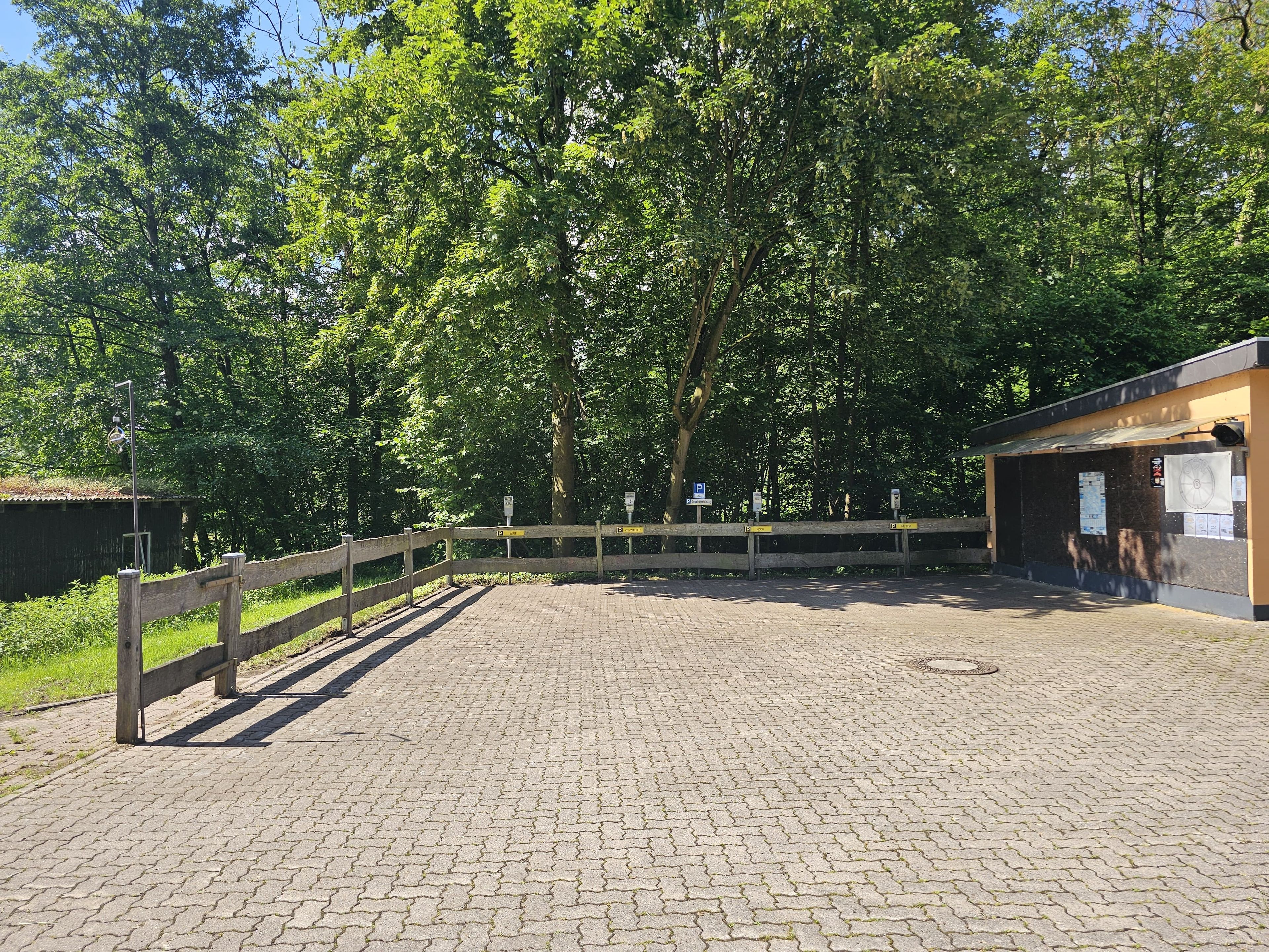 A paved parking area with a wooden fence, a small building with a sign, and trees in the background.