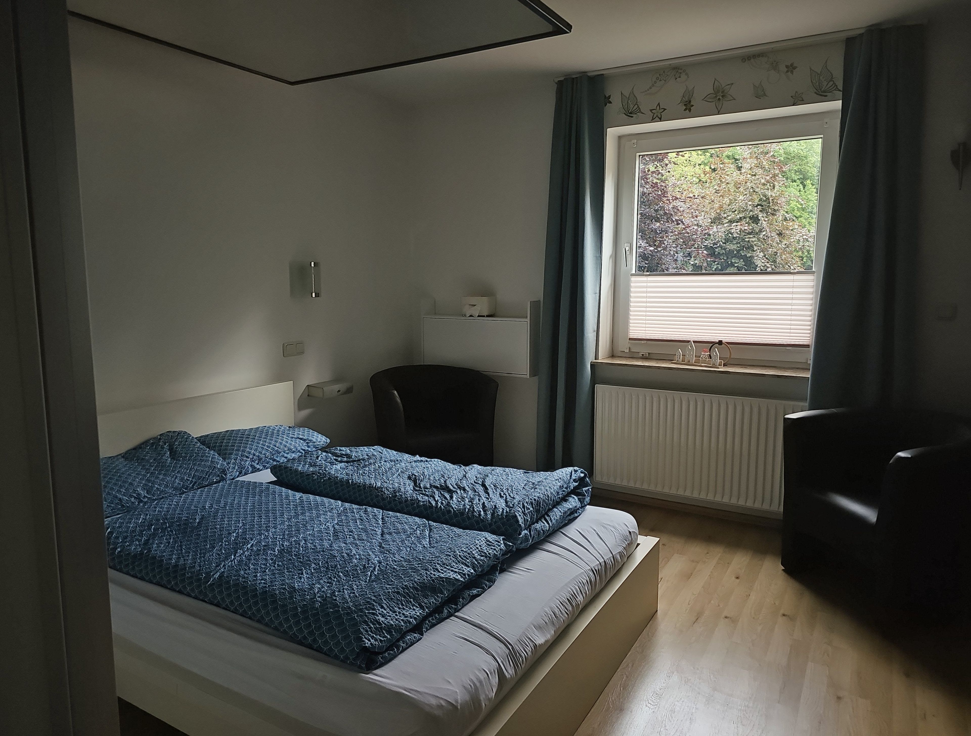 Ein Schlafzimmer mit einem Bett, einem Schreibtisch, einem Sessel, einem Fenster mit Vorhängen und einer Heizung.