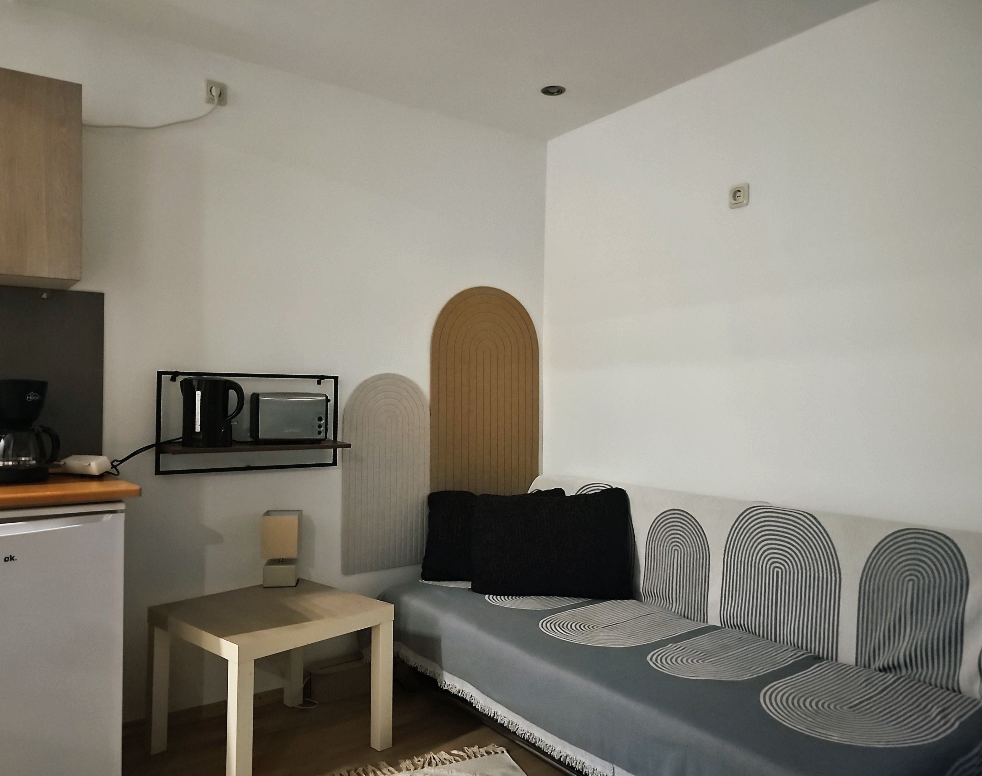 A compact living space with a kitchenette, a small table, a sofa with striped cushions, and a wall-mounted shelf with a kettle and a microwave.