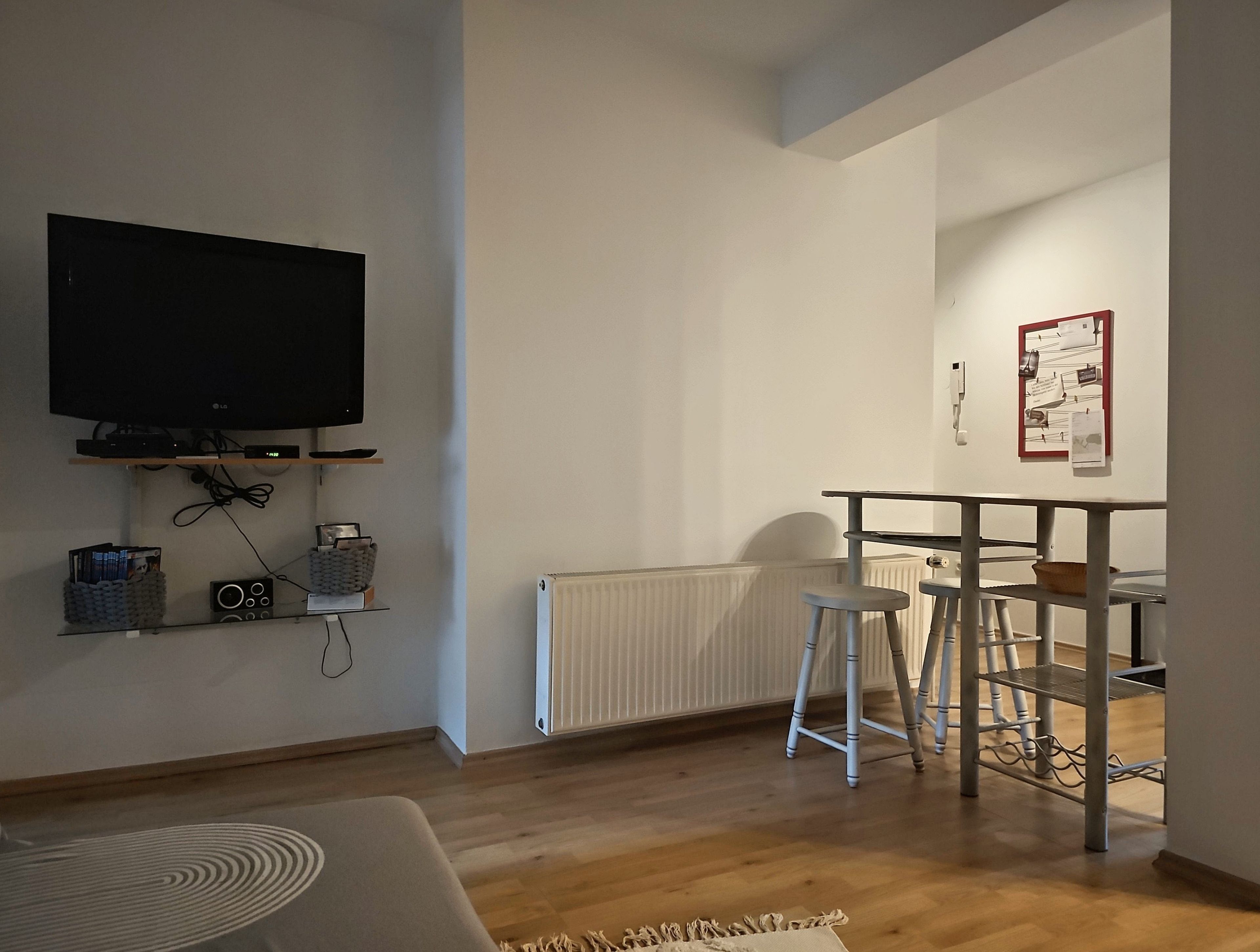 Ein modernes Wohnzimmer mit Flachbildfernseher, Heizkörper, Essbereich mit Barhocker und einem an der Wand befestigten Korkpinnbrett.