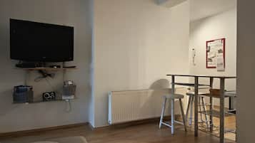 Ein modernes Wohnzimmer mit Flachbildfernseher, Heizkörper, Essbereich mit Barhocker und einem an der Wand befestigten Korkpinnbrett.