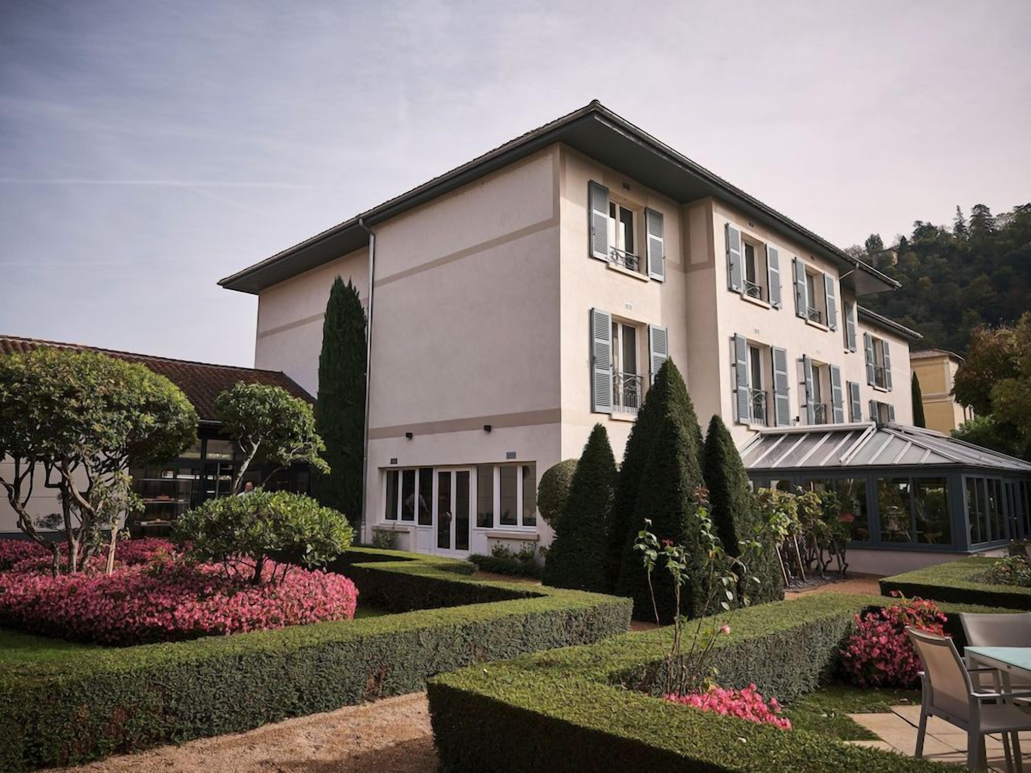 Un grand bâtiment à deux étages, doté d’une façade symétrique, d’une véranda et d’un jardin bien entretenu, agrémenté de haies et de plantes à fleurs.