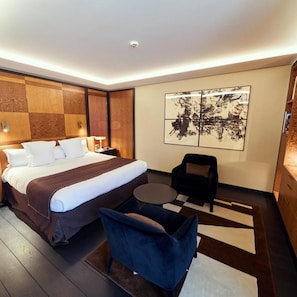 Una habitación de hotel moderna con una cama grande, una mesita redonda pequeña, un sillón azul y un cuadro abstracto de gran tamaño en la pared.