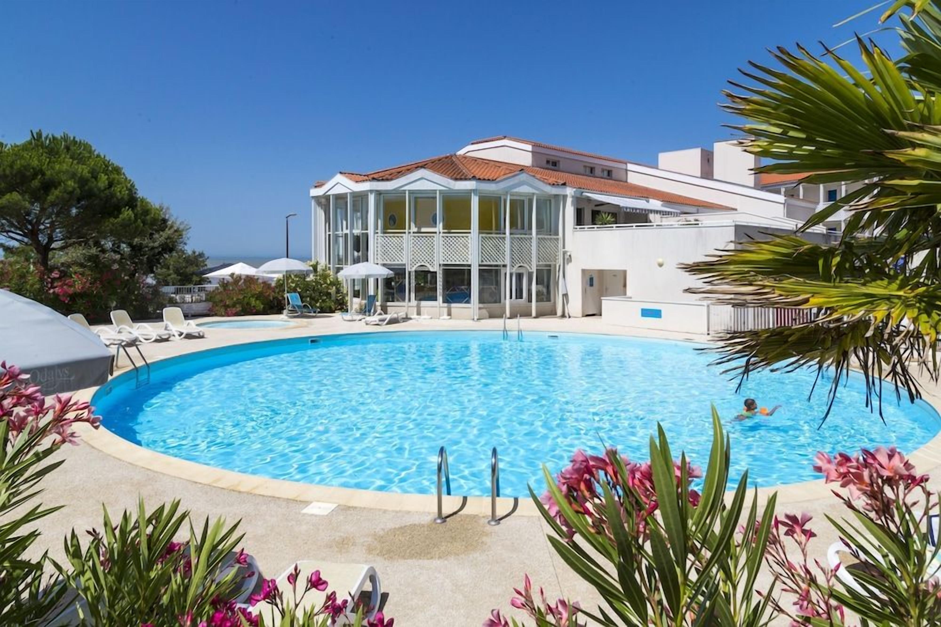 Une piscine entourée de chaises longues et de parasols, le tout bordé par un bâtiment à la façade blanche et au toit rouge.