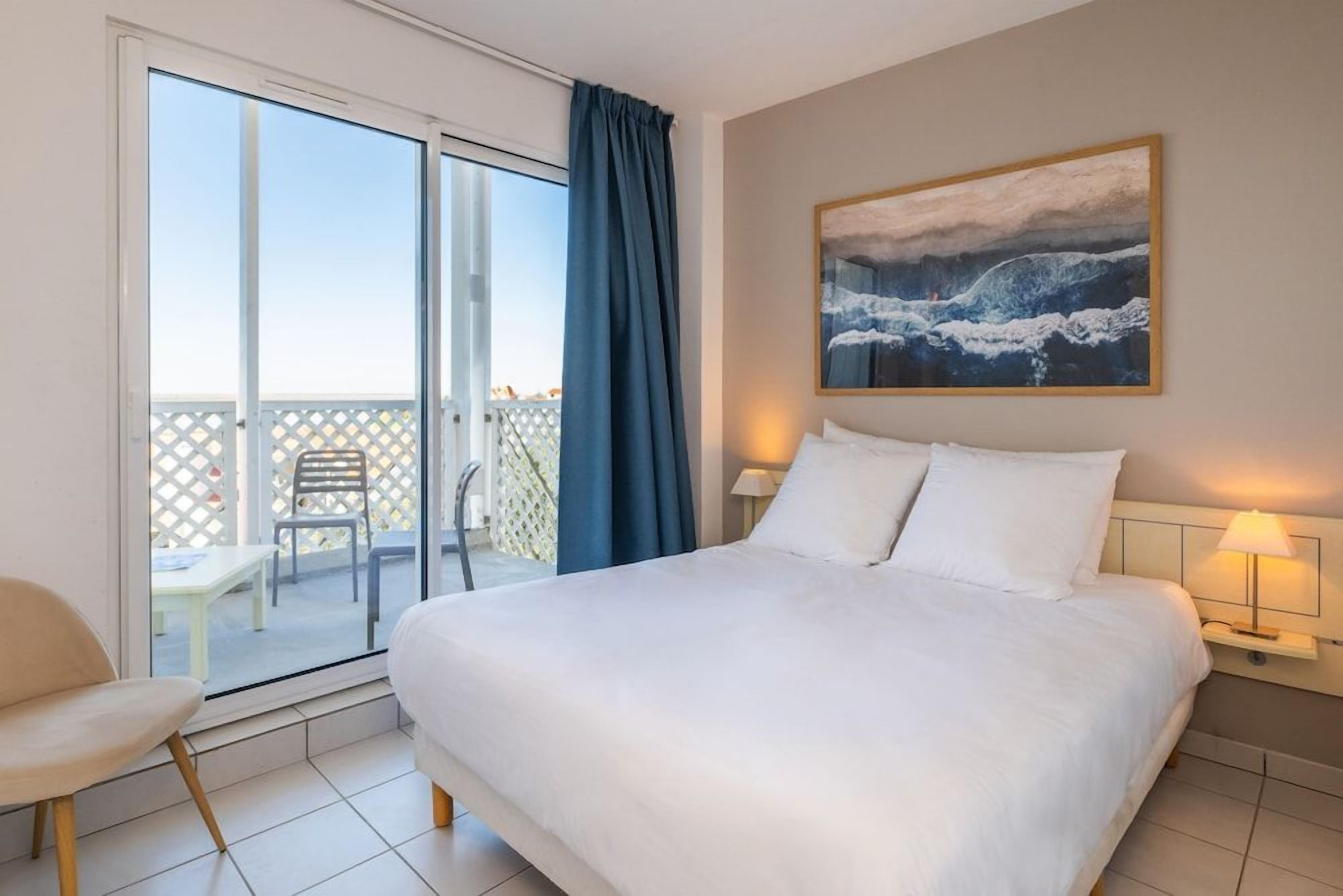 Une chambre d’hôtel avec un lit, une table de chevet avec une lampe, un tableau au mur et une porte-fenêtre coulissante donnant sur un espace extérieur avec une table et des chaises.