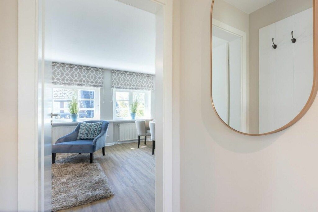A room with a round mirror, a chair, a sofa, and a view of a window with blinds.