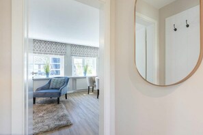 Ein Zimmer mit einem runden Spiegel, einem Sessel, einer Couch und Blick auf ein Fenster mit Jalousien.