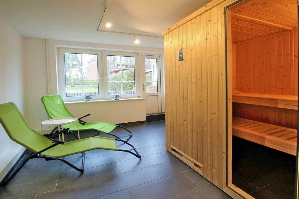 A room with two green lounge chairs, a window with a view of a house, and a sauna with wooden walls.