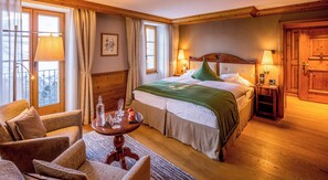 Ein Hotelzimmer mit einem Bett, zwei Nachttischen, einem Holzkopfende, einem Fenster mit Vorhängen, einem kleinen Tisch mit einer Lampe und einem Sessel.
