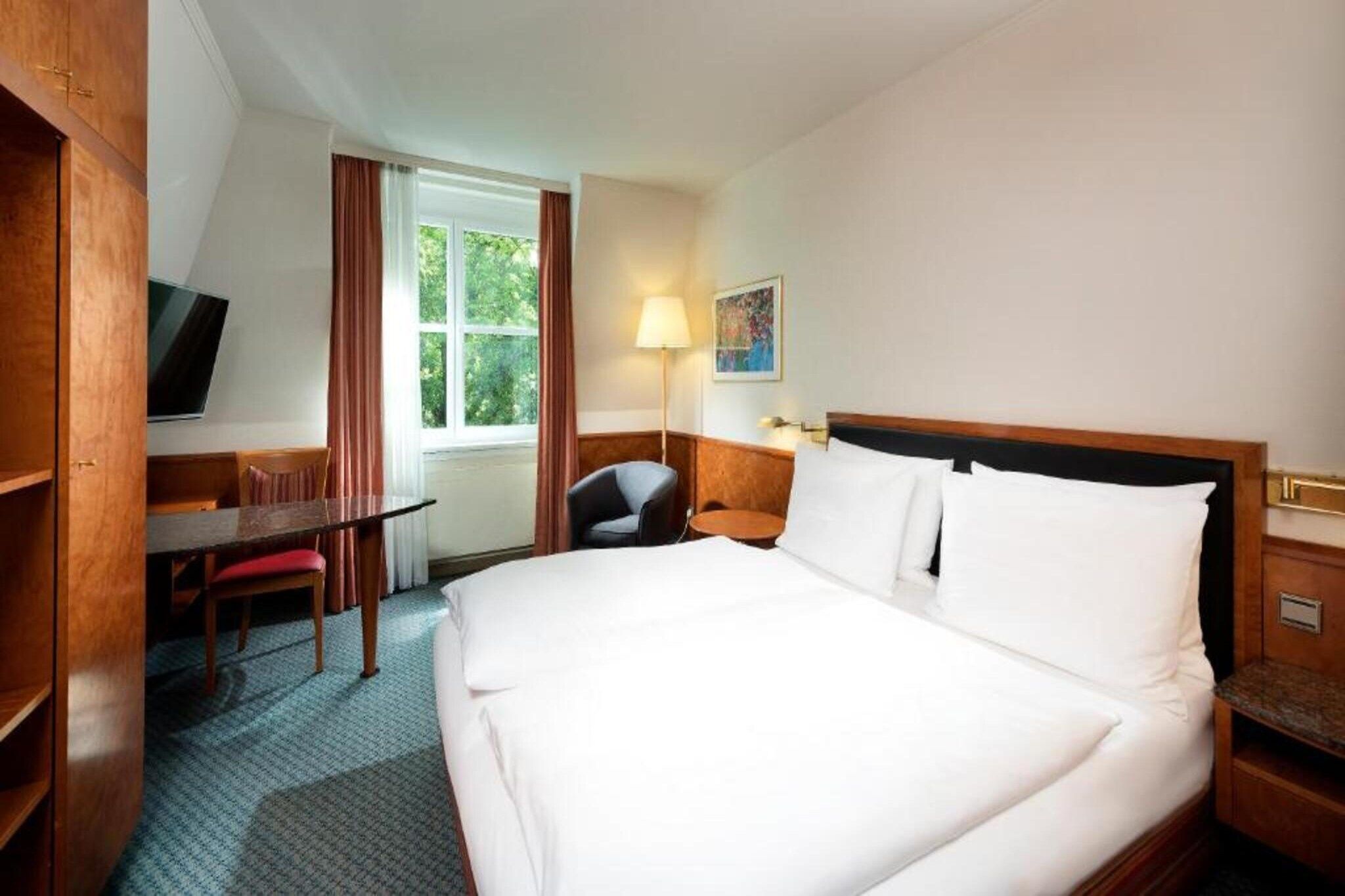 Ein Hotelzimmer mit einem großen Bett, einem Schreibtisch, einem Sessel, einem Fernseher und einem Fenster mit Vorhängen.