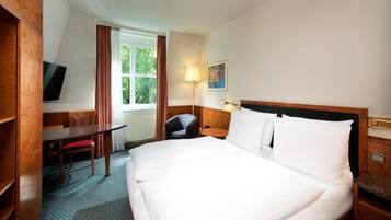 Ein Hotelzimmer mit einem großen Bett, einem Schreibtisch, einem Sessel, einem Fernseher und einem Fenster mit Vorhängen.