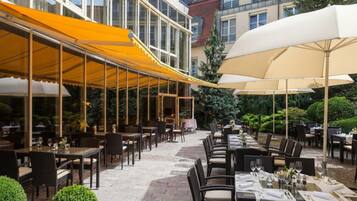An outdoor dining area with tables and chairs, covered by yellow and white canopies, and surrounded by greenery.