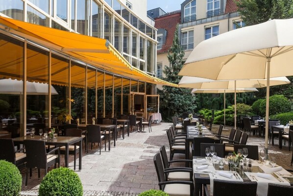 An outdoor dining area with tables and chairs, covered by yellow and white canopies, and surrounded by greenery.