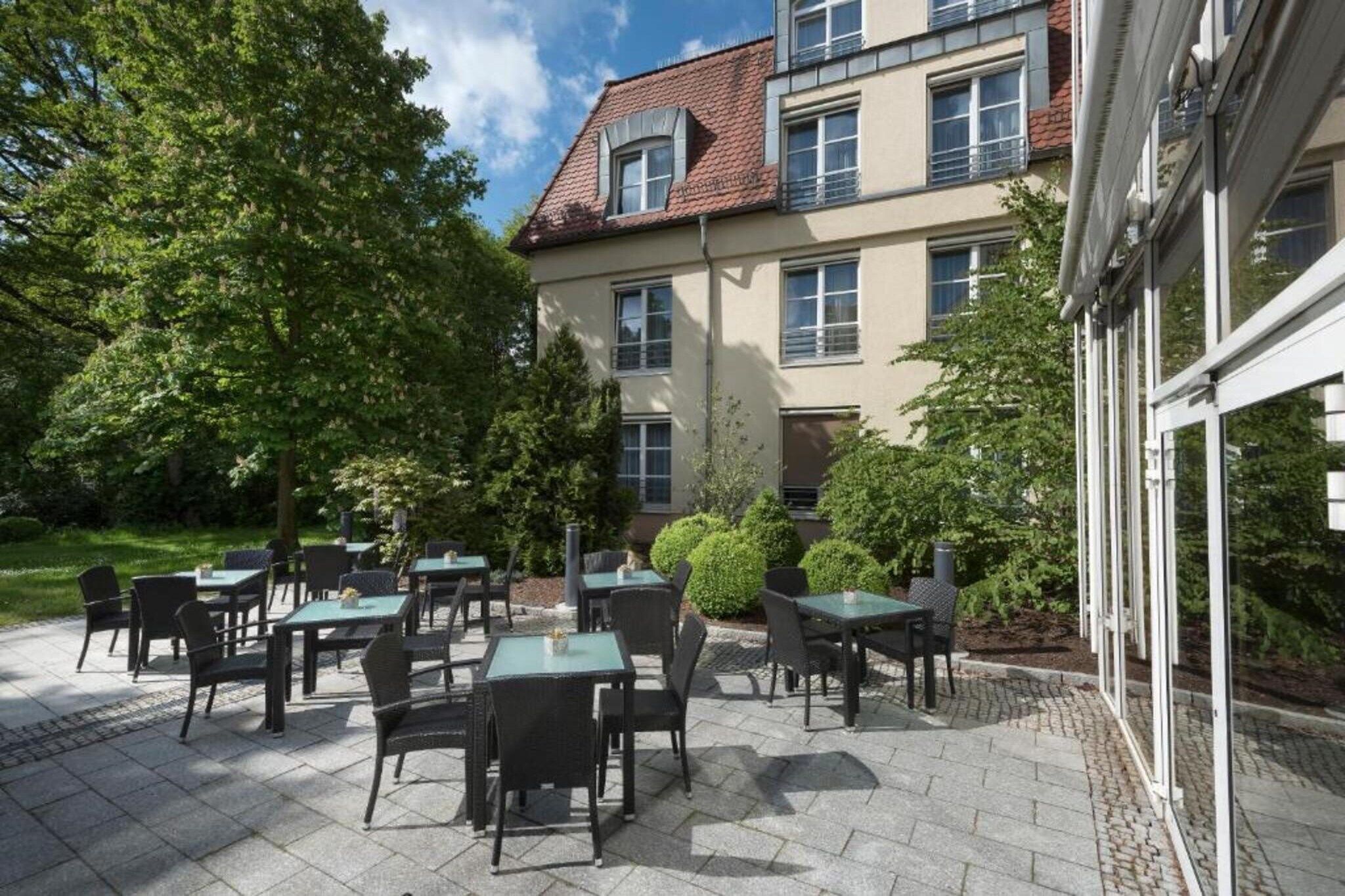 Un patio al aire libre con mesas y sillas, rodeado de árboles y un edificio con múltiples ventanales.