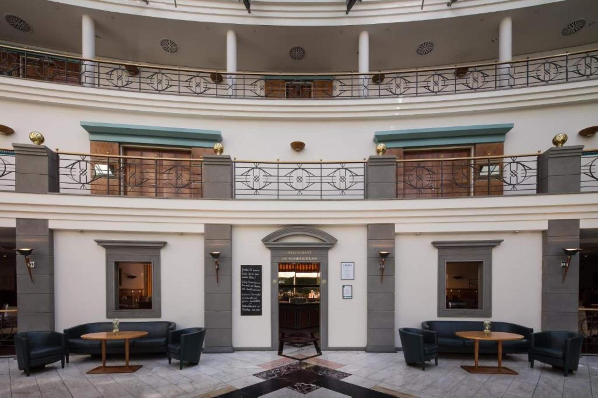 Eine mehrstöckige Hotelhalle mit einer zentralen Treppe, Sitzbereichen und Eingang zum Restaurant.