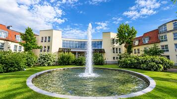 Ein runder Brunnen mit Wasserstrahlen in der Mitte, umgeben von einer gepflegten Rasenfläche und einem Gebäude mit zahlreichen Fenstern und Balkonen.