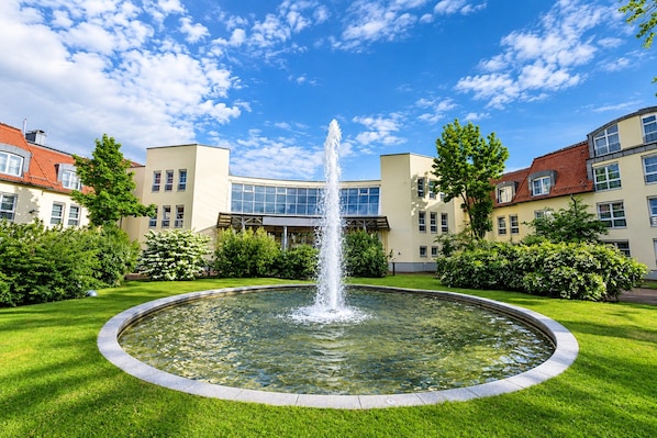 A circular fountain with water jets in the center, surrounded by a well-maintained lawn and a building with multiple windows and balconies. - Hotel Leipzig Superior Double Room (Leipzig)