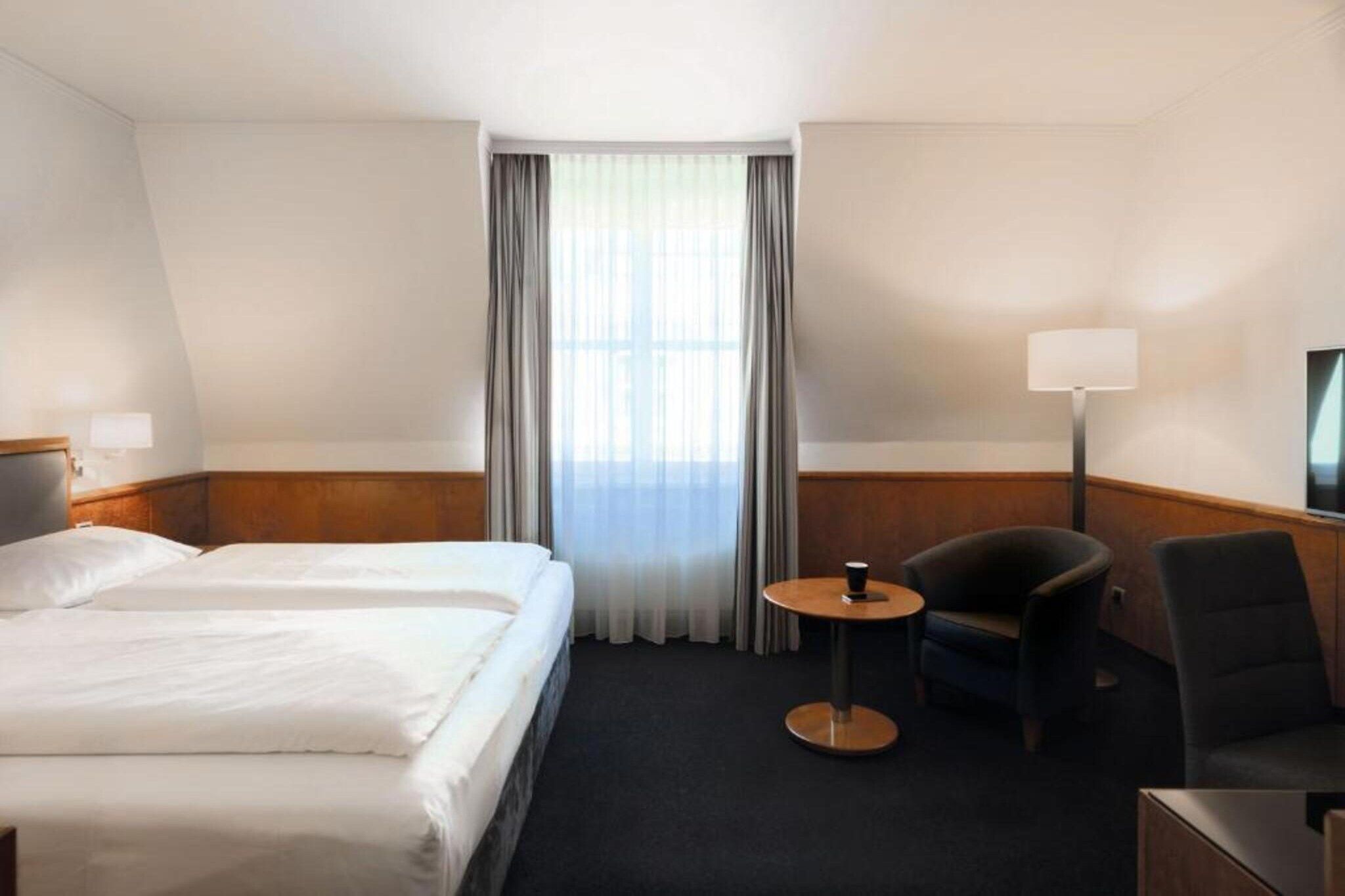 Ein Hotelzimmer mit zwei Betten, einem kleinen runden Tisch, einem Sessel, einem Fernseher und einem Fenster mit Vorhängen.