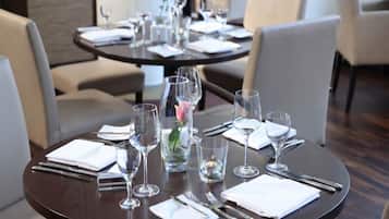 Une table à manger dressée avec des serviettes blanches, des couverts et de la vaisselle transparente, notamment des verres à vin et des verres à eau, dans un intérieur moderne agrémenté de chaises et d’un vase avec une fleur.