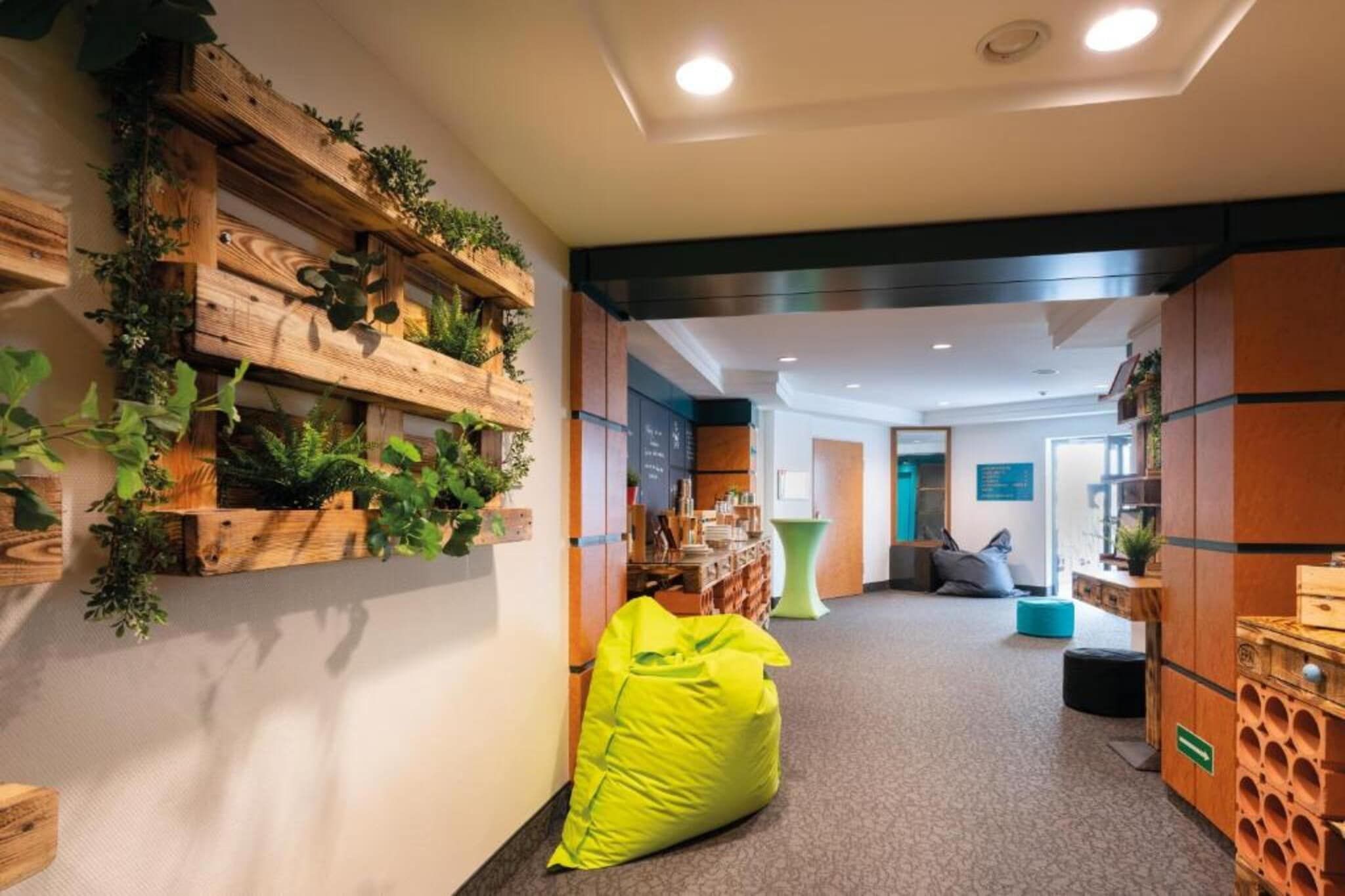 A hallway with a wooden wall adorned with greenery, a bright yellow bean bag chair, and a seating area with a blue cushion.