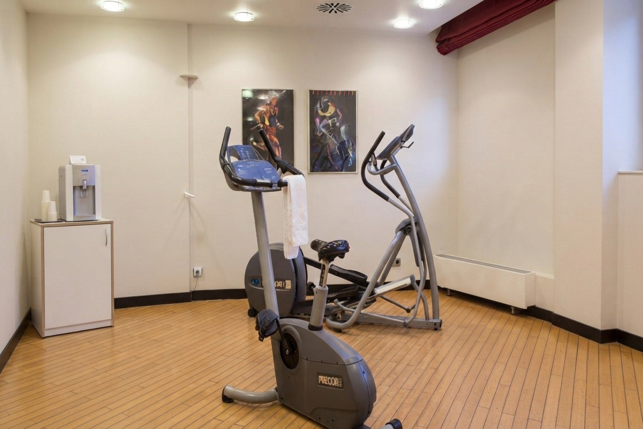 Ein Fitnessraum mit Trainingsgeräten, einem Wasserspender und zwei Wandplakaten.