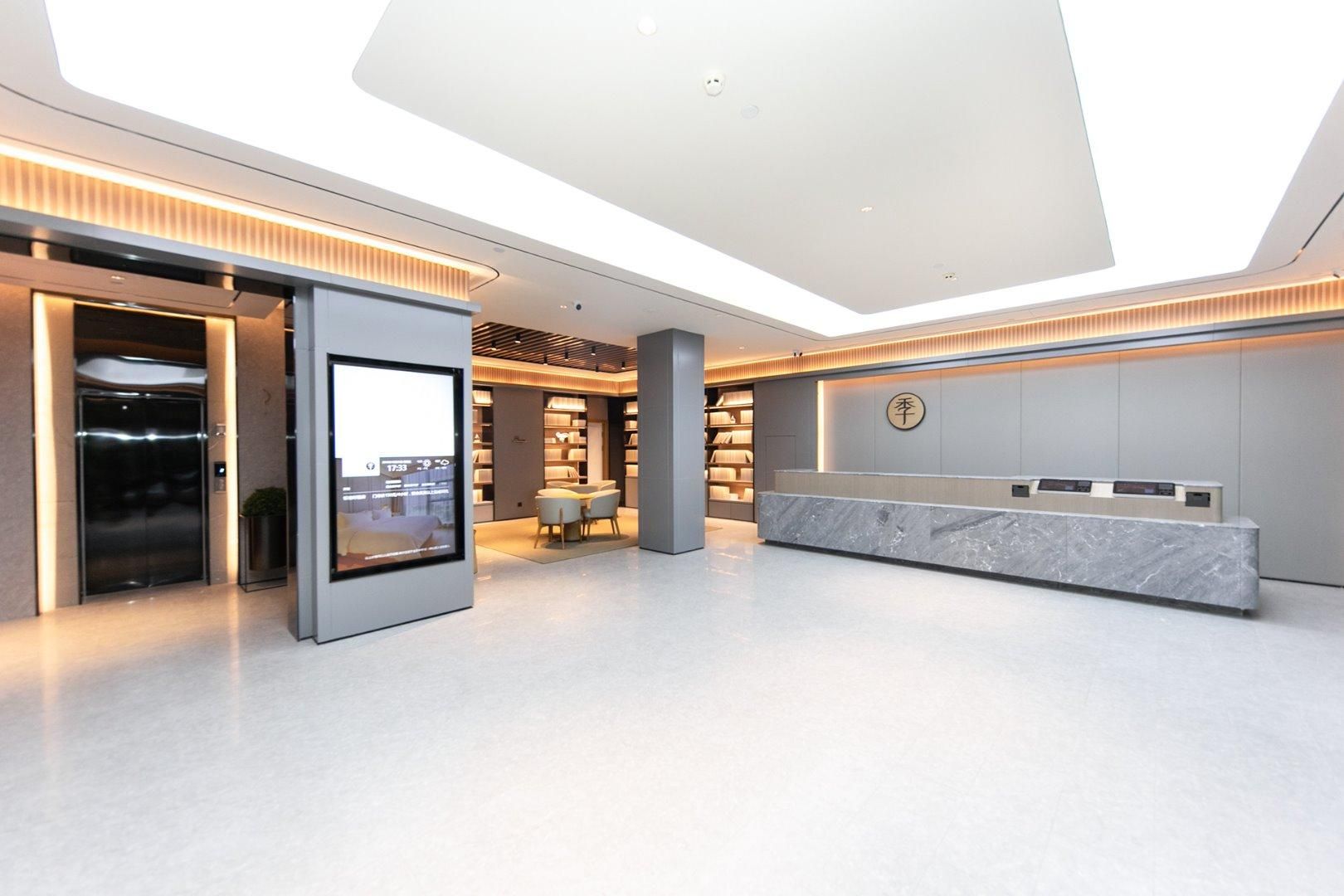 A modern hotel lobby with a reception desk, a digital display, a seating area, and a bookshelf.