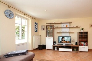 A living room with a television, a sofa, a clock, and a window. - Lüttmarsch 13, Holiday Apartment 2 (Wyk auf Föhr)