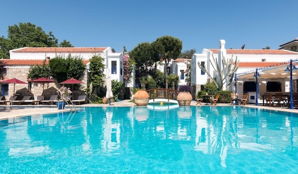 Una piscina con sillones de descanso y sombrillas, rodeada de edificios blancos con tejados de tejas rojas.