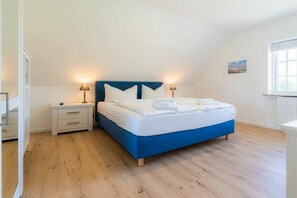 A bedroom with a blue bed, white bedding, two bedside tables with lamps, and a window with blinds. - Holiday apartment 7 "Langeneß" (ID 476) (Tating)