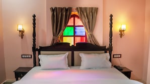 A double bed with white linens, two bedside tables with lamps, a colorful tapestry at the foot of the bed, and a window with stained glass. - Jhalra Heritage Haveli Jodhpur (Jodhpur)