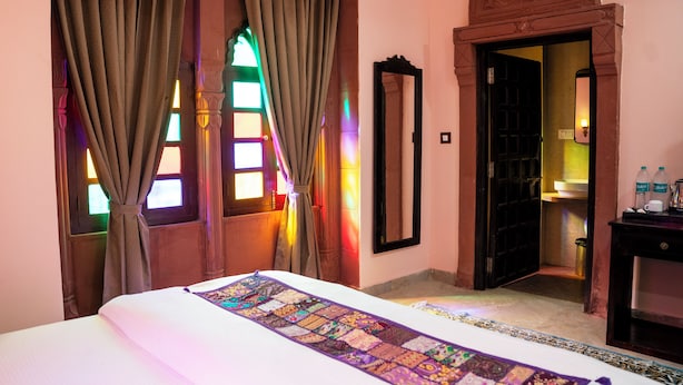 A hotel room with a bed, curtains, a mirror, a door, and a small table with bottles. - Jhalra Heritage Haveli Jodhpur (Jodhpur)