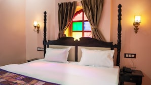 A bed with a headboard, two bedside tables, a wall-mounted lamp, and a window with curtains. - Jhalra Heritage Haveli Jodhpur (Jodhpur)