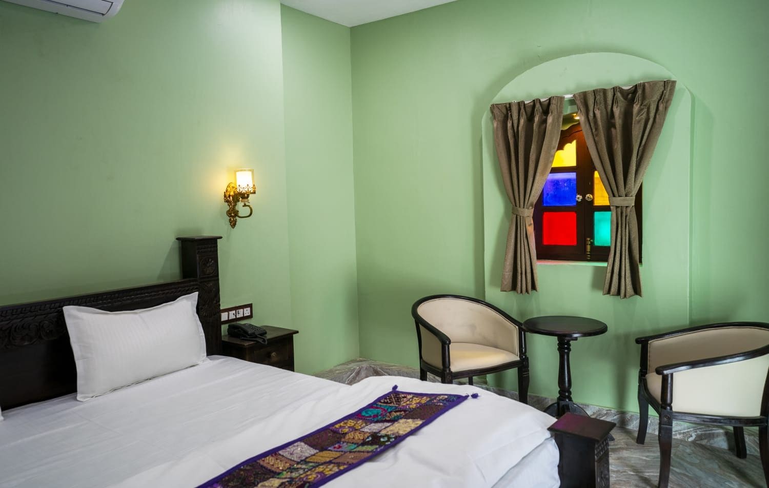 Habitación de hotel con cama, dos sillas, una mesita, una mesita de noche, una lámpara de pared y un ventanal con cortinas.