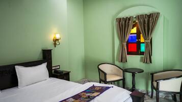 Habitación de hotel con cama, dos sillas, una mesita, una mesita de noche, una lámpara de pared y un ventanal con cortinas.